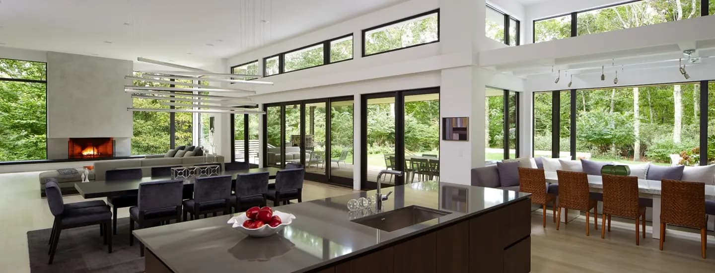 An open-concept great room with floor-to-ceiling black-framed Andersen windows and gliding patio doors overlooking the woods