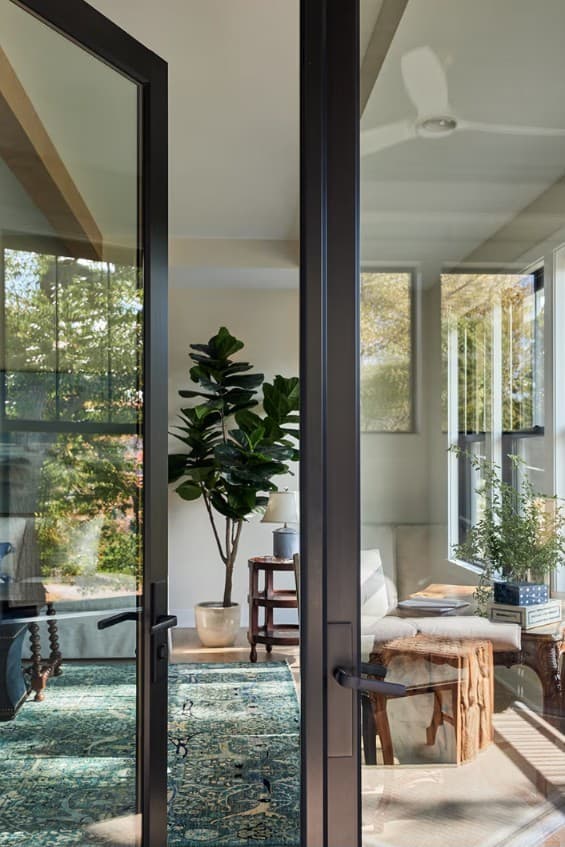 A bright sunroom seen through open black-framed glass doors, with a fiddle-leaf fig and coastal furnishings