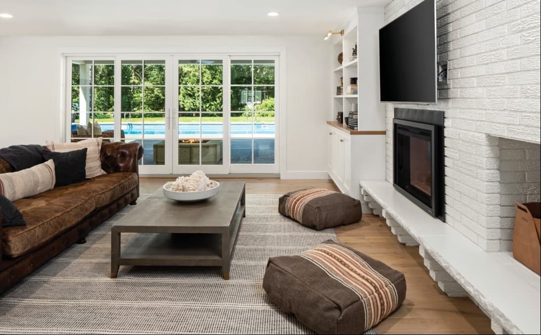 A living room with white-framed sliding glass doors opening to a pool, beside a white-brick fireplace