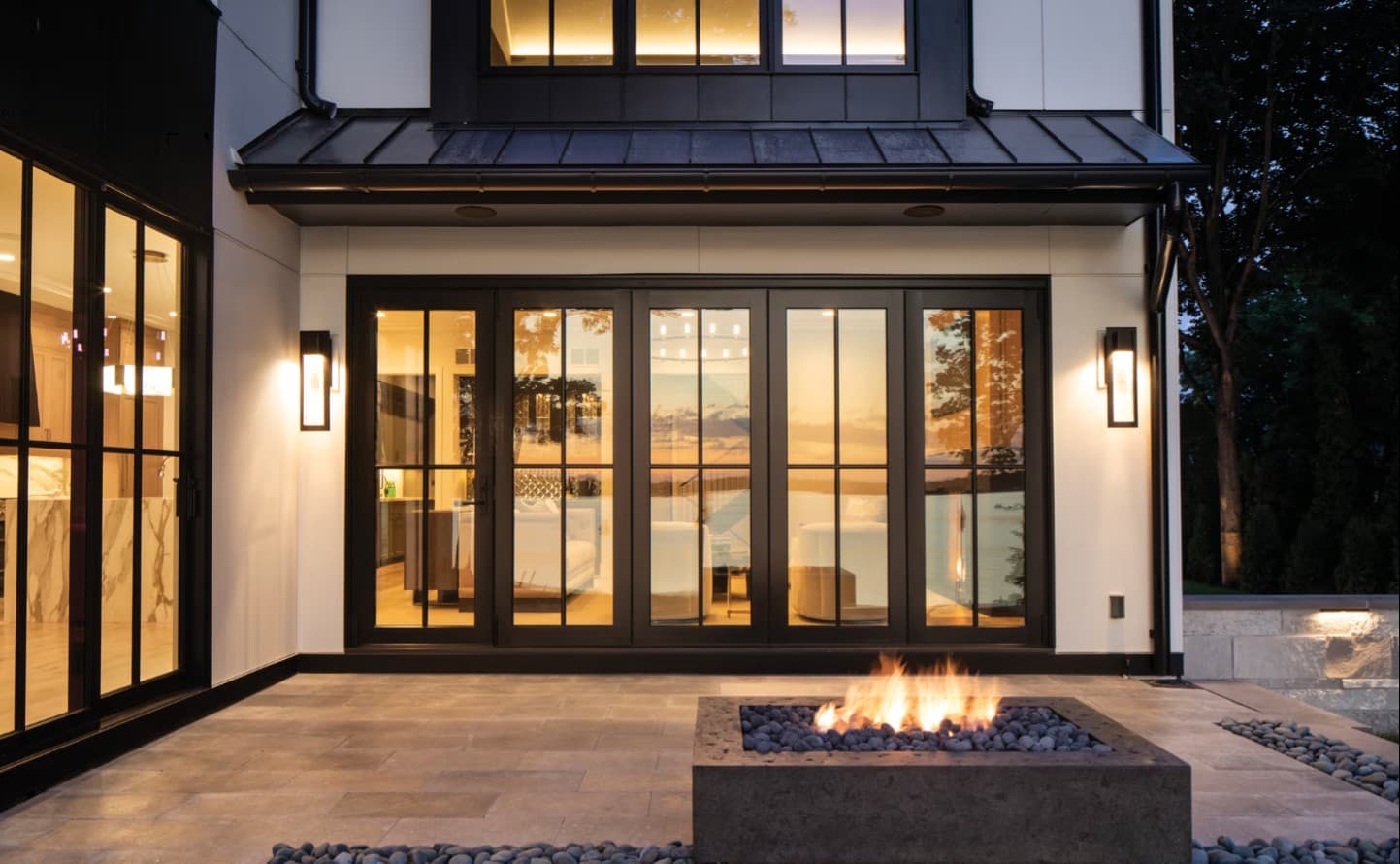 A modern farmhouse at dusk with a wall of black-framed folding glass doors glowing warm above a fire-pit patio by the water