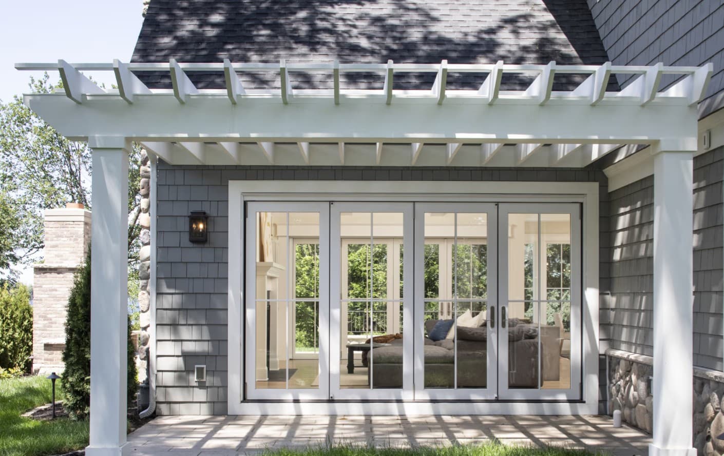 A gray shingle-sided coastal home with a pergola over a patio and white-framed sliding glass doors to the living room