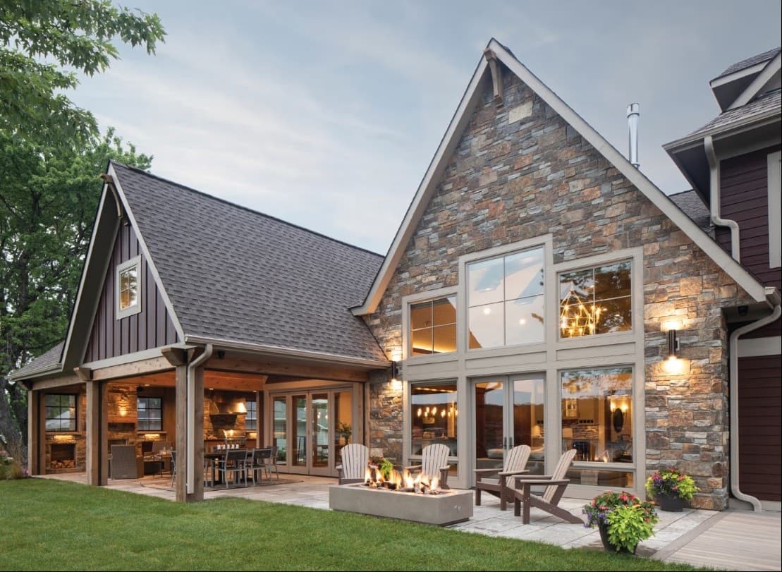 A luxury modern farmhouse at dusk with board-and-batten and stone gable ends, large black-grid windows, French doors, and a covered porch beside a fire-pit patio