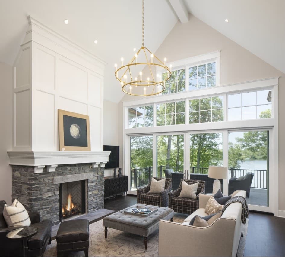 A coastal great room with a two-story wall of white grid windows and doors overlooking a lake, anchored by a stone fireplace