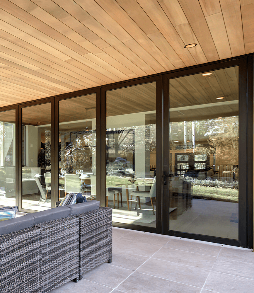 Wide black-framed panoramic sliding glass doors on a covered patio, opening a dining room to the outdoors under a wood-plank ceiling