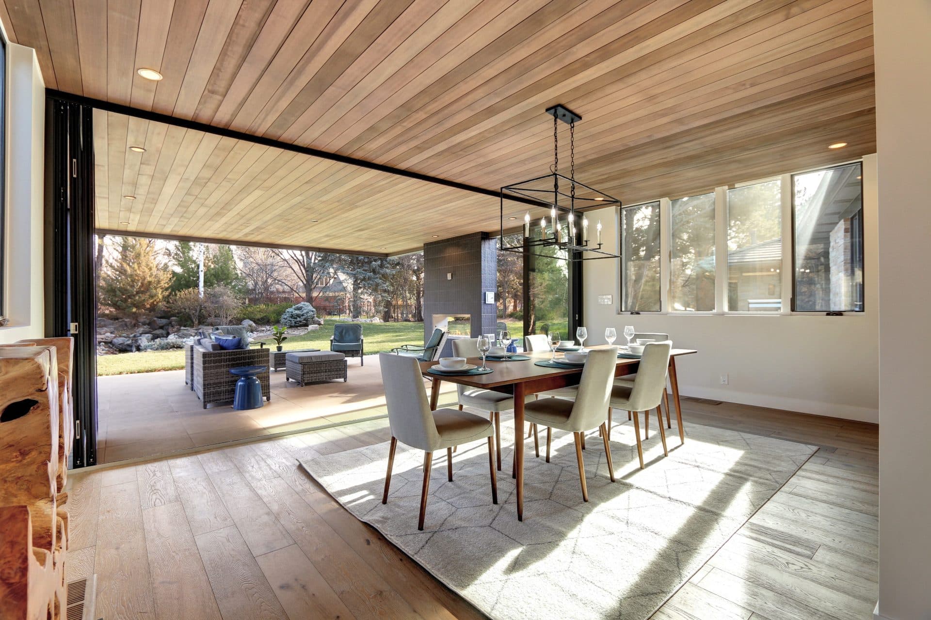 A dining room with a wall of black bi-fold doors folded fully open to a patio under a wood-plank ceiling