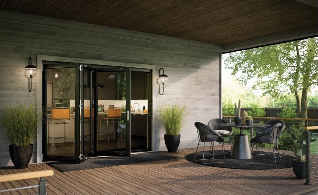 A covered deck with black-framed bi-fold glass doors folded open to a modern kitchen, beside a dining set