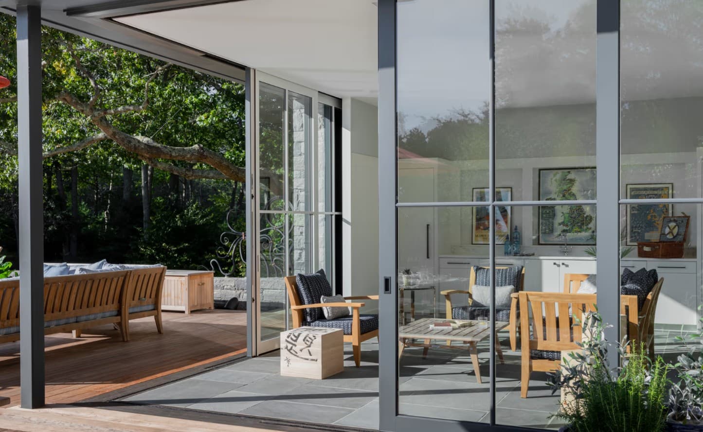 A covered bluestone patio with tall black-framed sliding glass doors beside a mature oak, opening to a sitting room