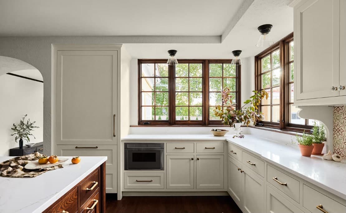 A bright white kitchen with warm wood-framed Marvin divided-light windows above marble countertops