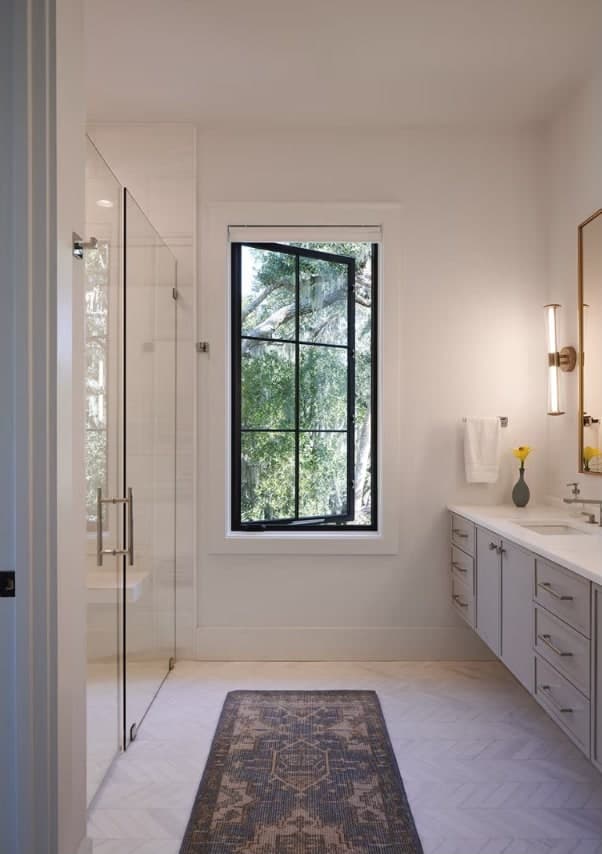 A tall black-framed divided-light casement window over a herringbone floor in a bright white bathroom