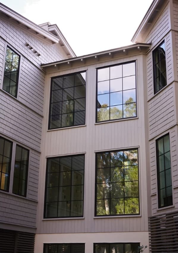 The exterior of a shingle-sided coastal home with tall black-framed Marvin grid windows wrapping a corner, framed by trees