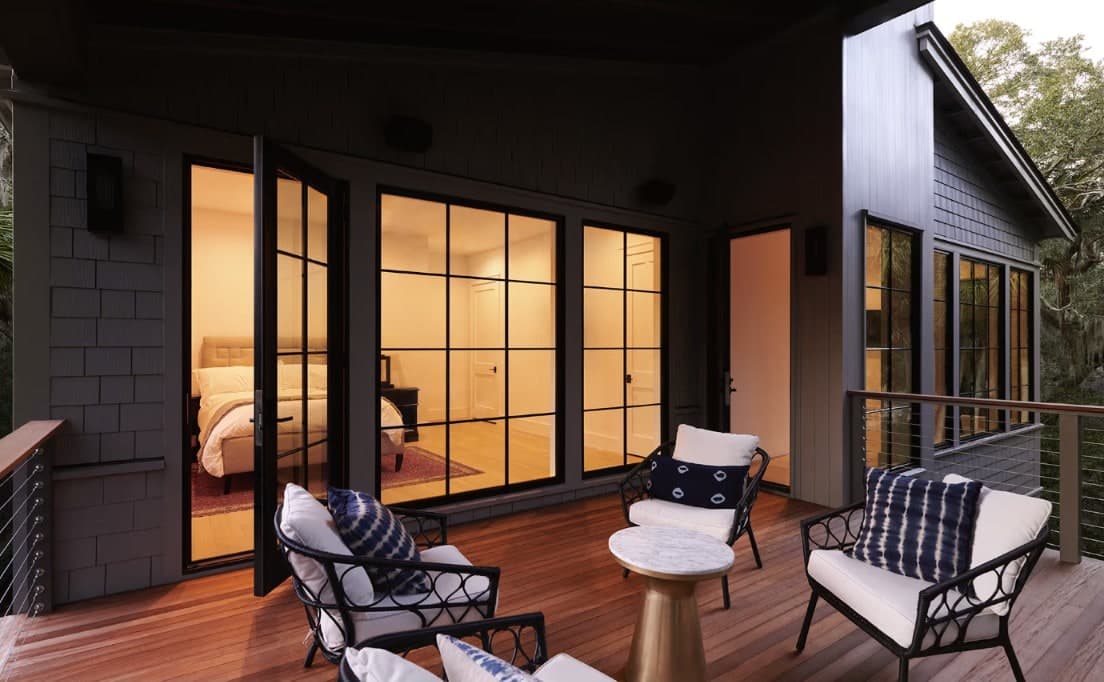 Black-framed French doors and windows glowing warm at dusk on a covered coastal deck with lounge seating