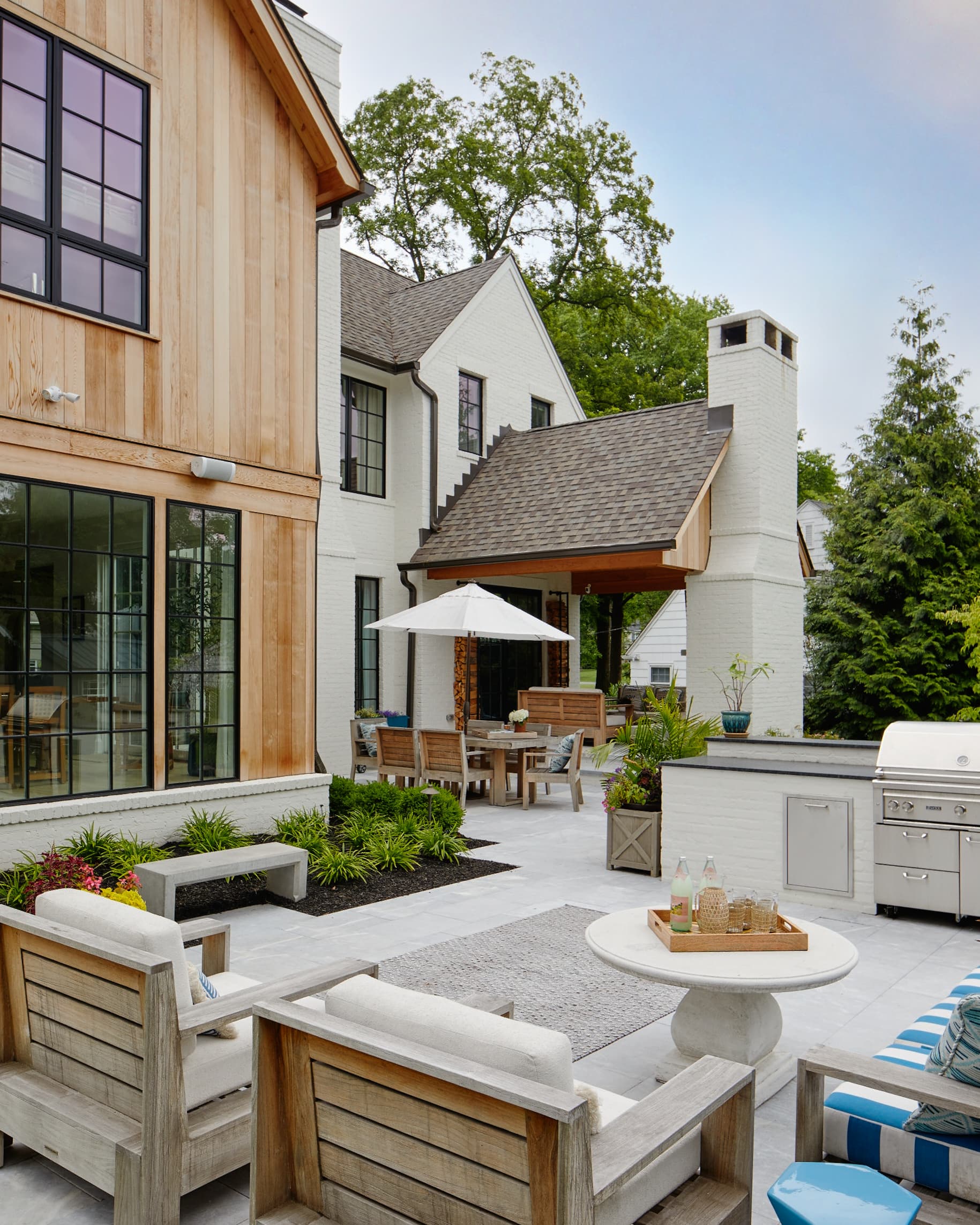 A modern farmhouse with natural cedar and white-brick siding, large black-grid windows, and an outdoor-kitchen patio