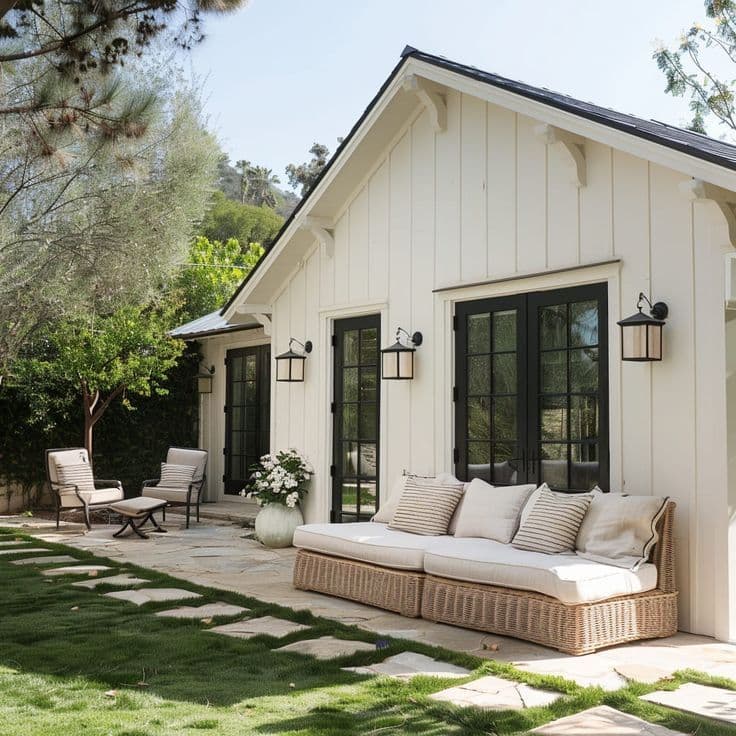 A white board-and-batten guest cottage with black-framed French doors and windows, black lantern sconces, and a wicker sofa on a flagstone patio