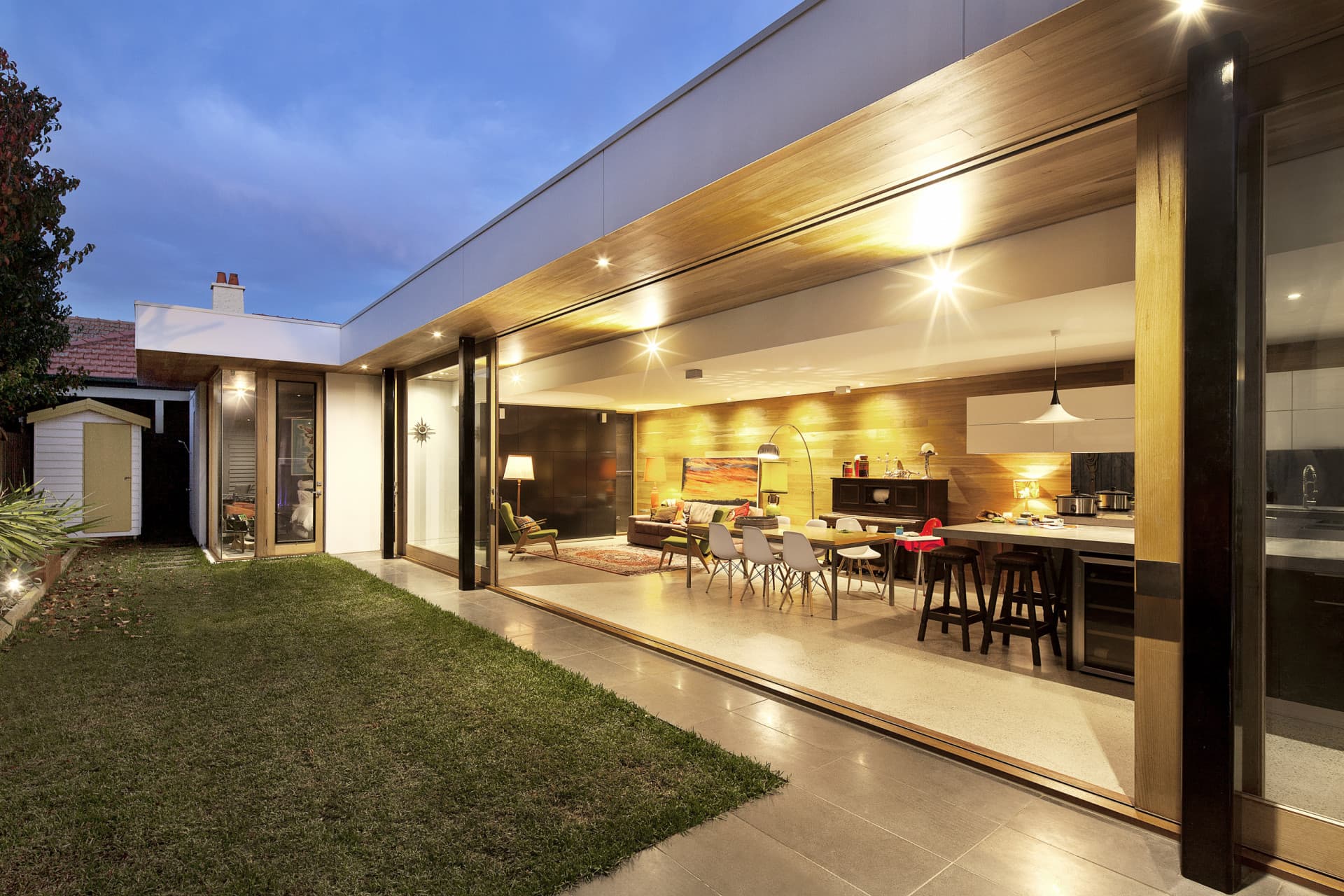 A modern home at dusk with a wide corner wall of sliding glass patio doors pocketed fully open, connecting an open-plan living and kitchen to a lawn and patio