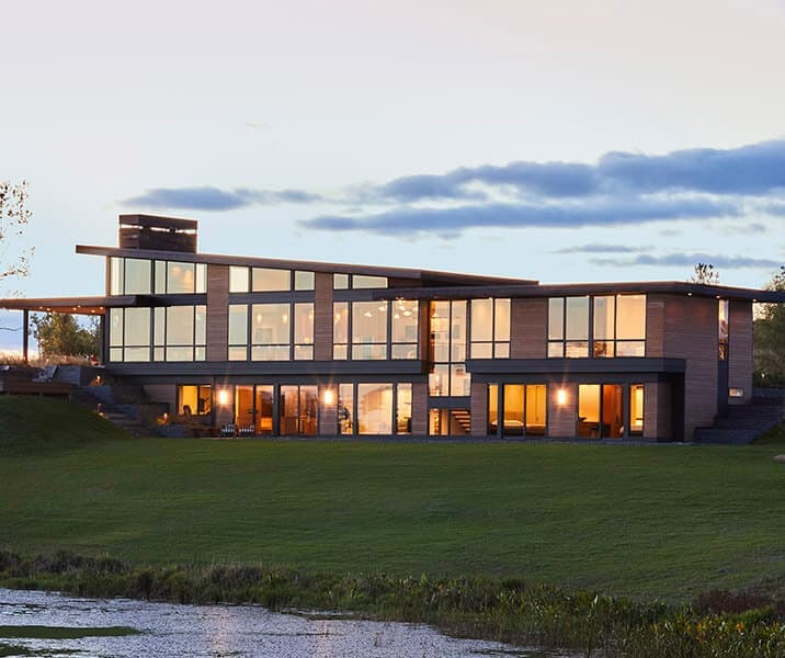 A modern waterfront home at dusk with walls of black-framed Pella windows glowing warm