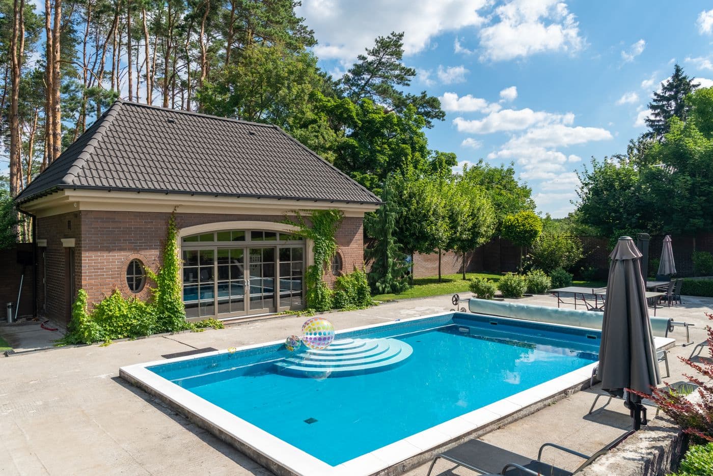 A detached brick pool house with arched glass French doors and ivy beside a rectangular pool