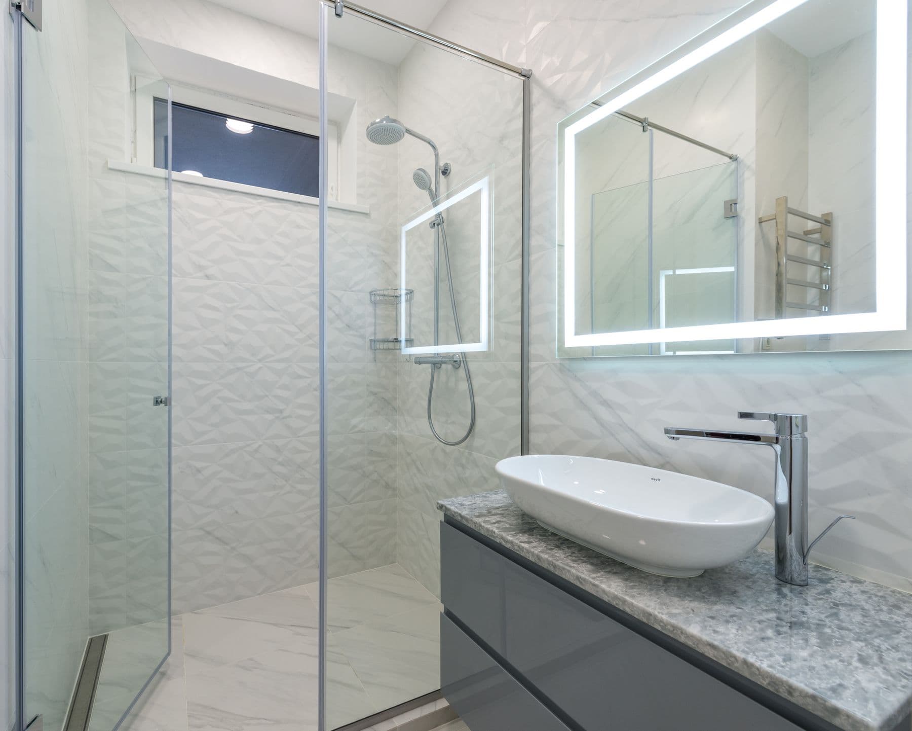 A modern bathroom with a glass walk-in shower, backlit mirror, and marble-look tile