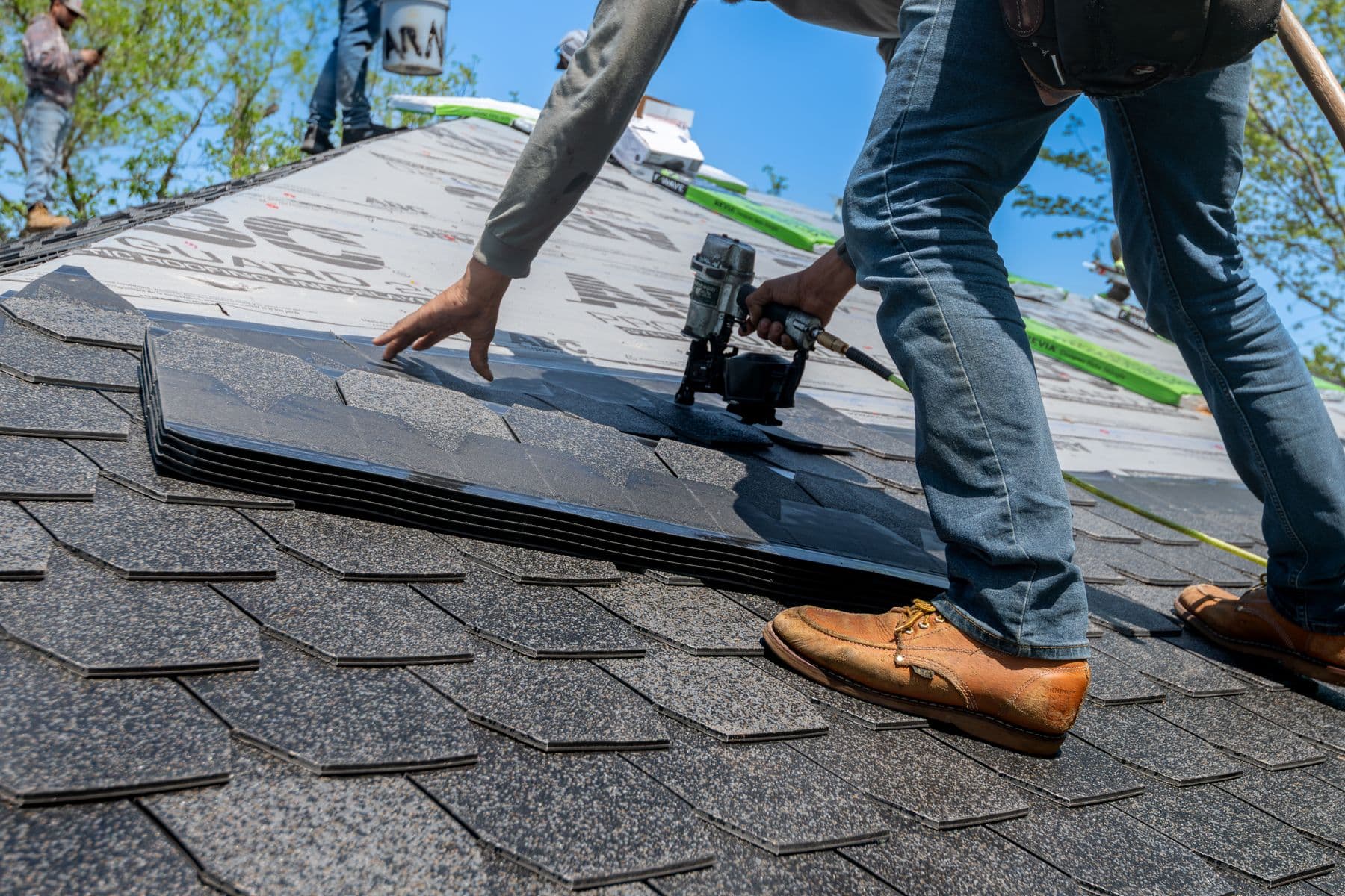 A roofer installing architectural asphalt shingles on a house roof under a blue sky