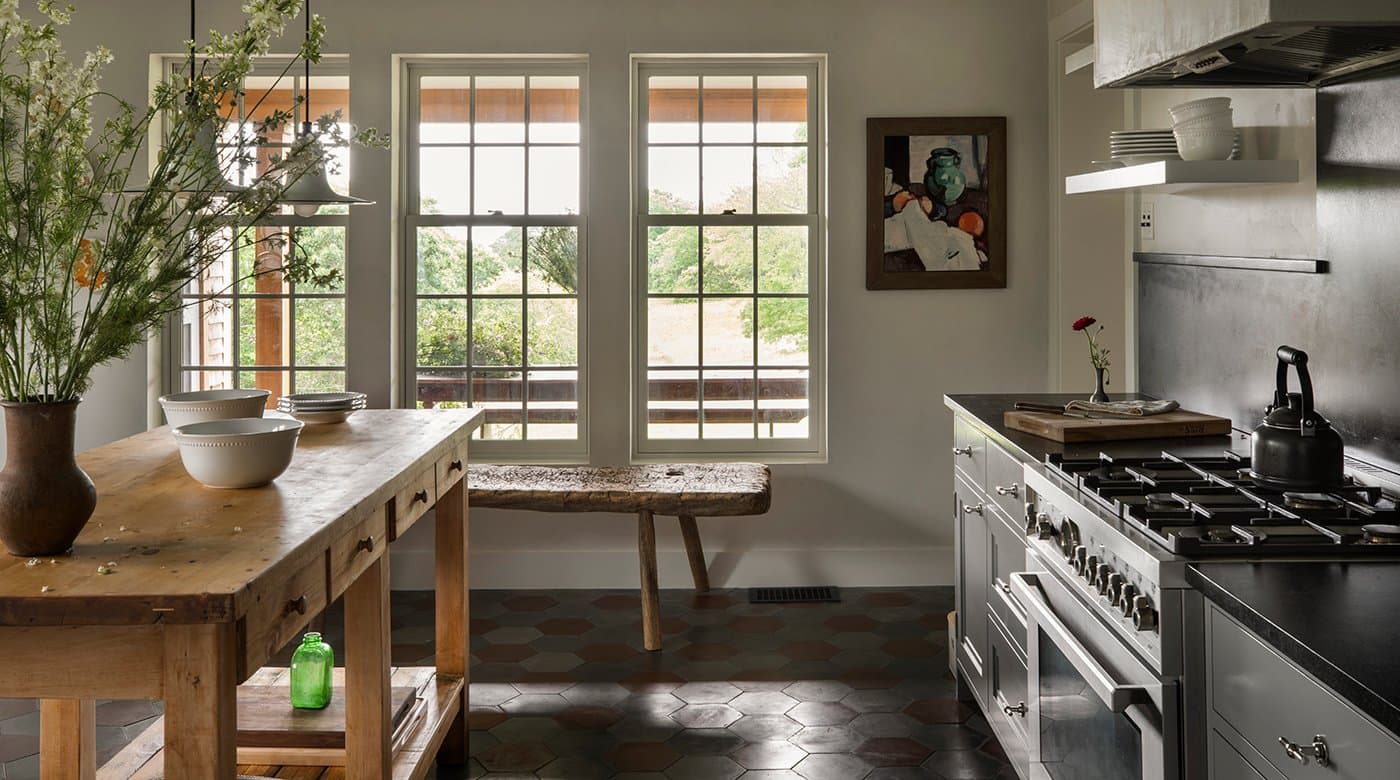 A farmhouse kitchen with tall white-framed Andersen double-hung windows with colonial grilles over a rustic wood table