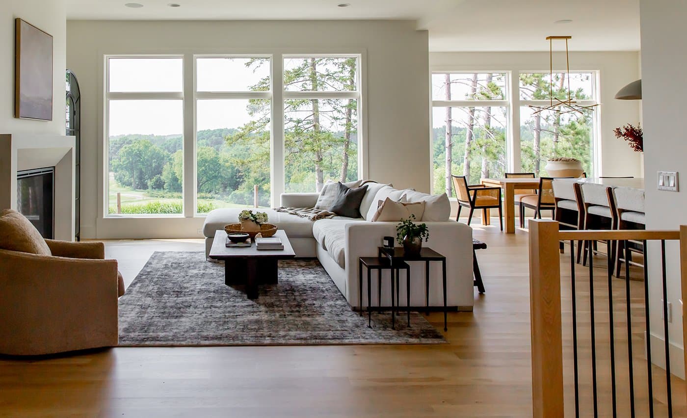 An open-concept living and dining room with a wall of white-framed Andersen picture and transom windows overlooking woods