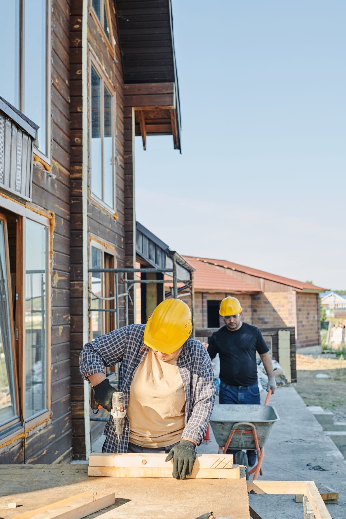 A construction worker drilling a board on a residential jobsite while a coworker moves materials behind