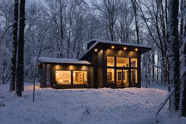 A dark modern cabin lit from within at dusk in the snow, clad in LP SmartSide