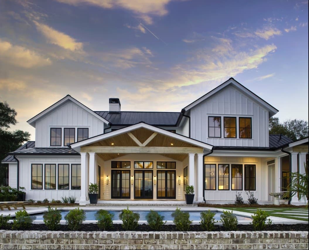 A white modern farmhouse with black-framed windows, a standing-seam metal roof, and a columned porch with black French doors mirrored in a reflecting pool at dusk